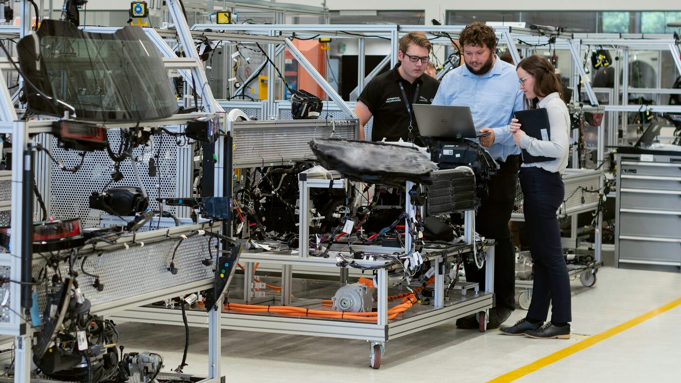 Three engineers at a laptop beside a test rig, with cabling and instrumentation exposed.